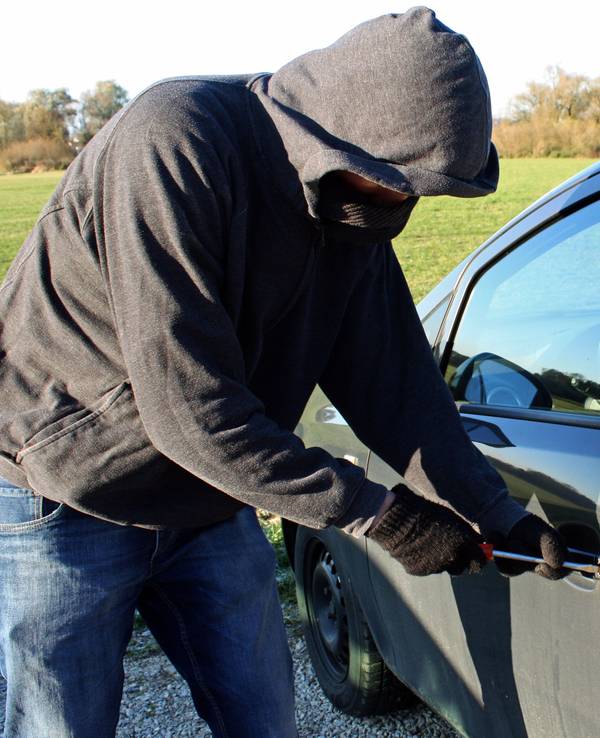 Ein vermummter Mann mit Kapuze bricht mit seinem Werkzeug ein Auto auf.