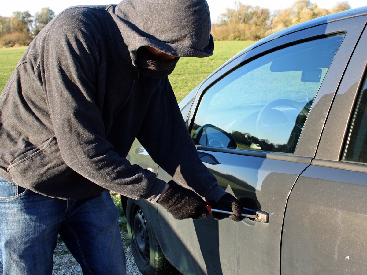Ein vermummter Mann mit Kapuze bricht mit seinem Werkzeug ein Auto auf.