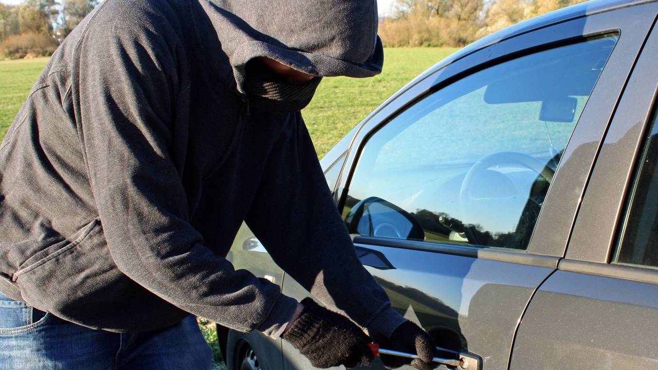 Ein vermummter Mann mit Kapuze bricht mit seinem Werkzeug ein Auto auf.