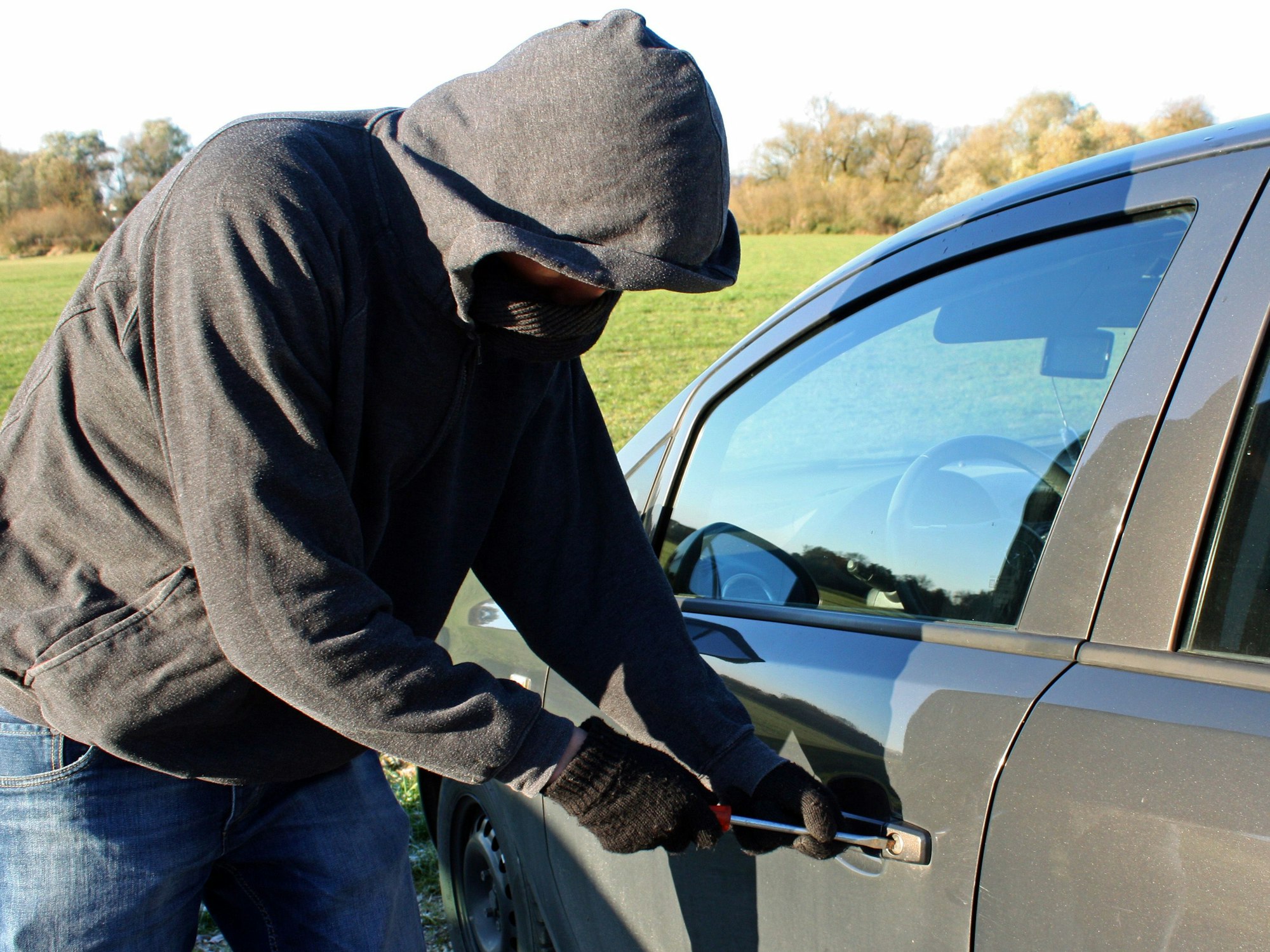 Ein vermummter Mann mit Kapuze bricht mit seinem Werkzeug ein Auto auf.