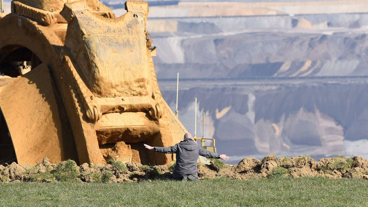 Ein Mann kniet vor einem Braunkohlebaggr und breitet die Arme aus.