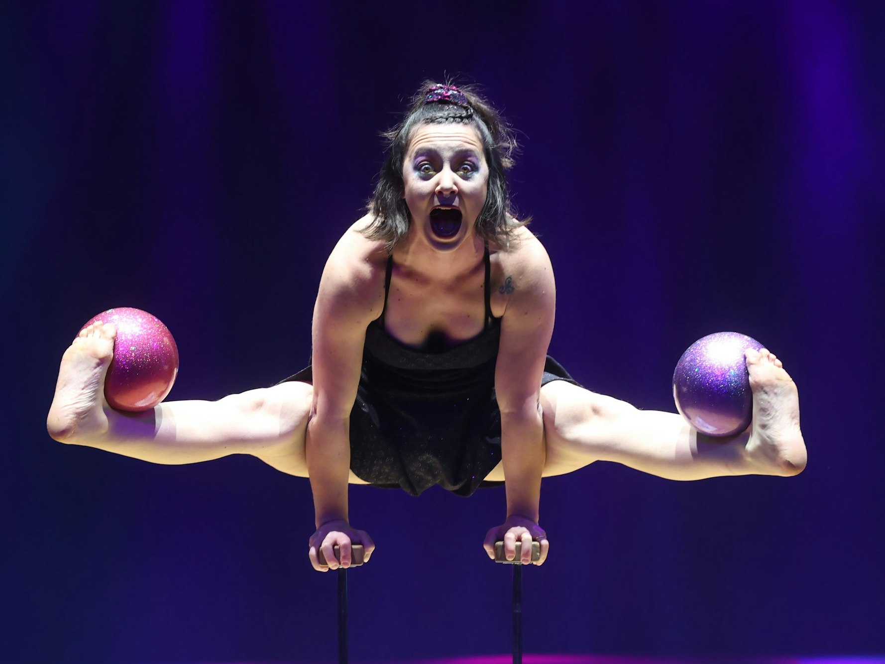 Weihnachtscircus-Artistin Sofi Poppisperatti stützt sich auf ihre Arme und spreizt ihre Beine. Zwischen ihren Füßen und ihren Schienbeinen hat sie Bowlingkugeln platziert. Ihr Mund ist aufgerissen.