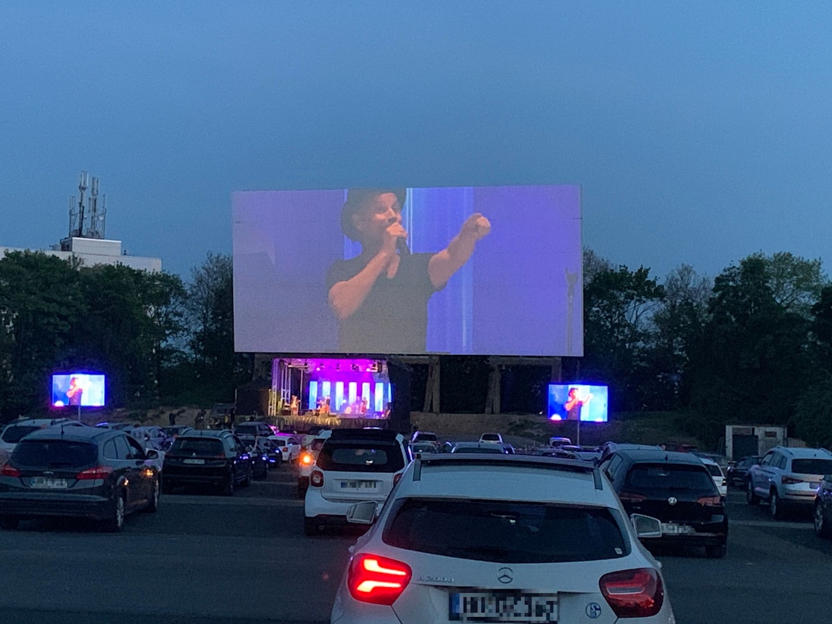 Dutzende Autos stehen vor der Bühne im Autokino in Köln-Porz. Dort steht die Band Brings auf der Bühne.