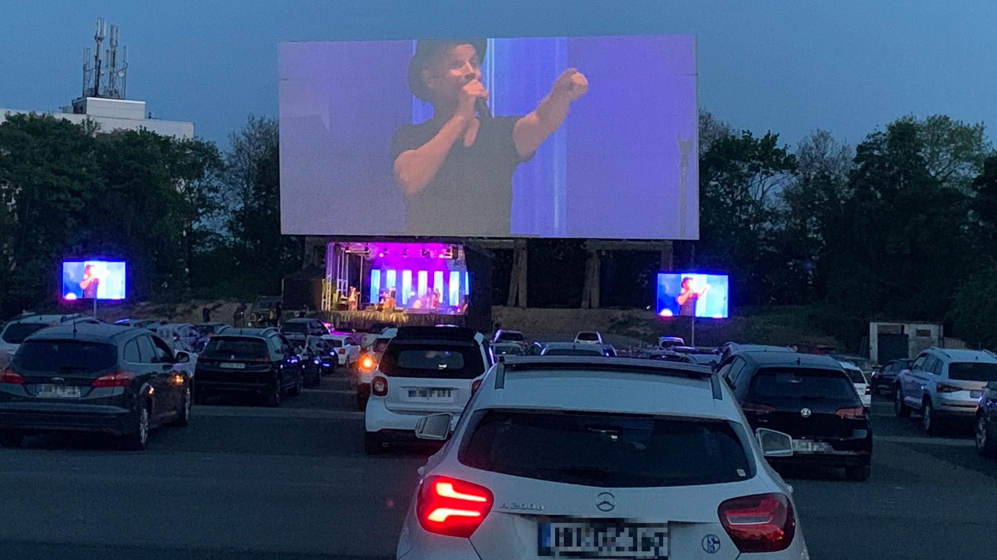Dutzende Autos stehen vor der Bühne im Autokino in Köln-Porz. Dort steht die Band Brings auf der Bühne.