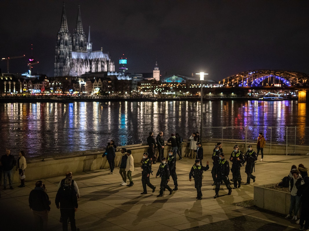 Menschen stehen am Rheinboulevard in Köln und schauen rüber auf die hell erleuchtete Zone rund um den Dom.