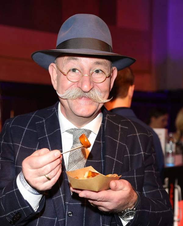 Horst Lichter lächelt beim Deutschen Fernsehpreis in einem Anzug in die Kamera und isst Pommes mit Currywurst.