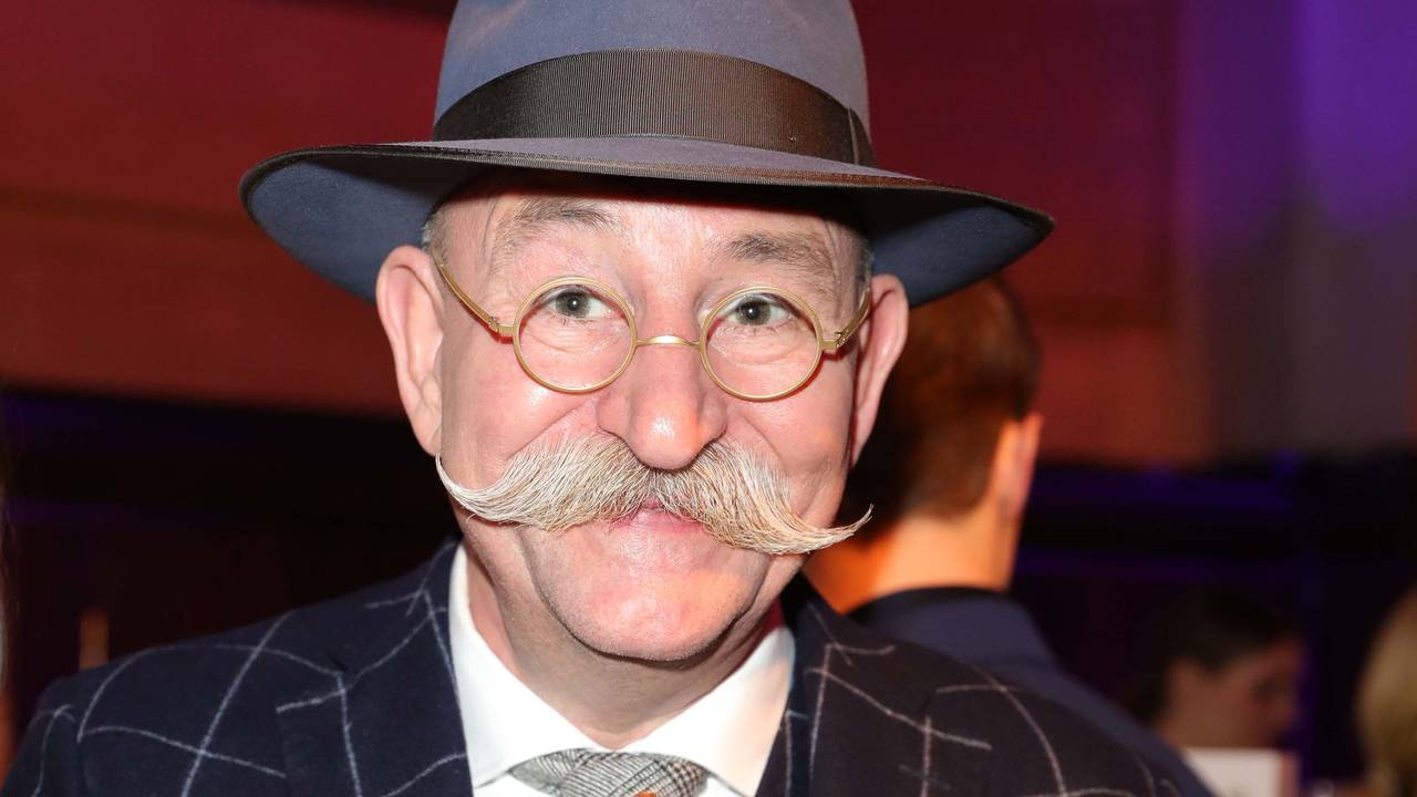 Horst Lichter lächelt beim Deutschen Fernsehpreis in einem Anzug in die Kamera und isst Pommes mit Currywurst.