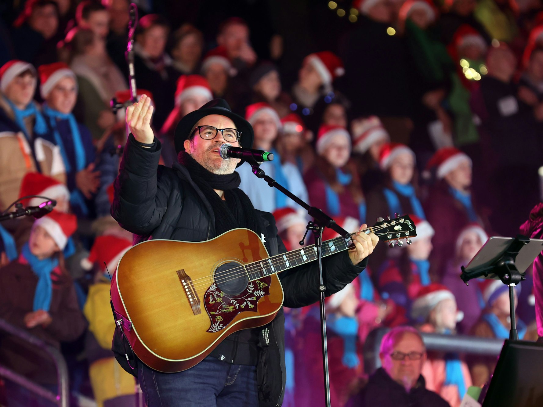 Ein Mann mit Gitarre steht vor zahlreichen Menschen, die eine Weihnachtsmann-Mütze tragen.