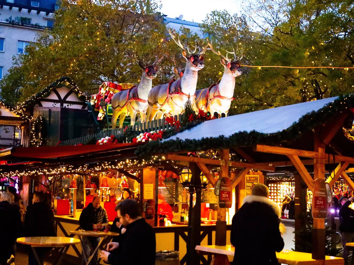 Menschen stehen an einem Weihnachtsmarktstand. Auf dem Dach stehen Rentiere, die einen Schlitten ziehen.