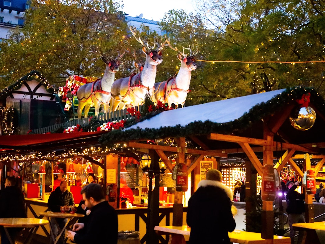 Menschen stehen an einem Weihnachtsmarktstand. Auf dem Dach stehen Rentiere, die einen Schlitten ziehen.