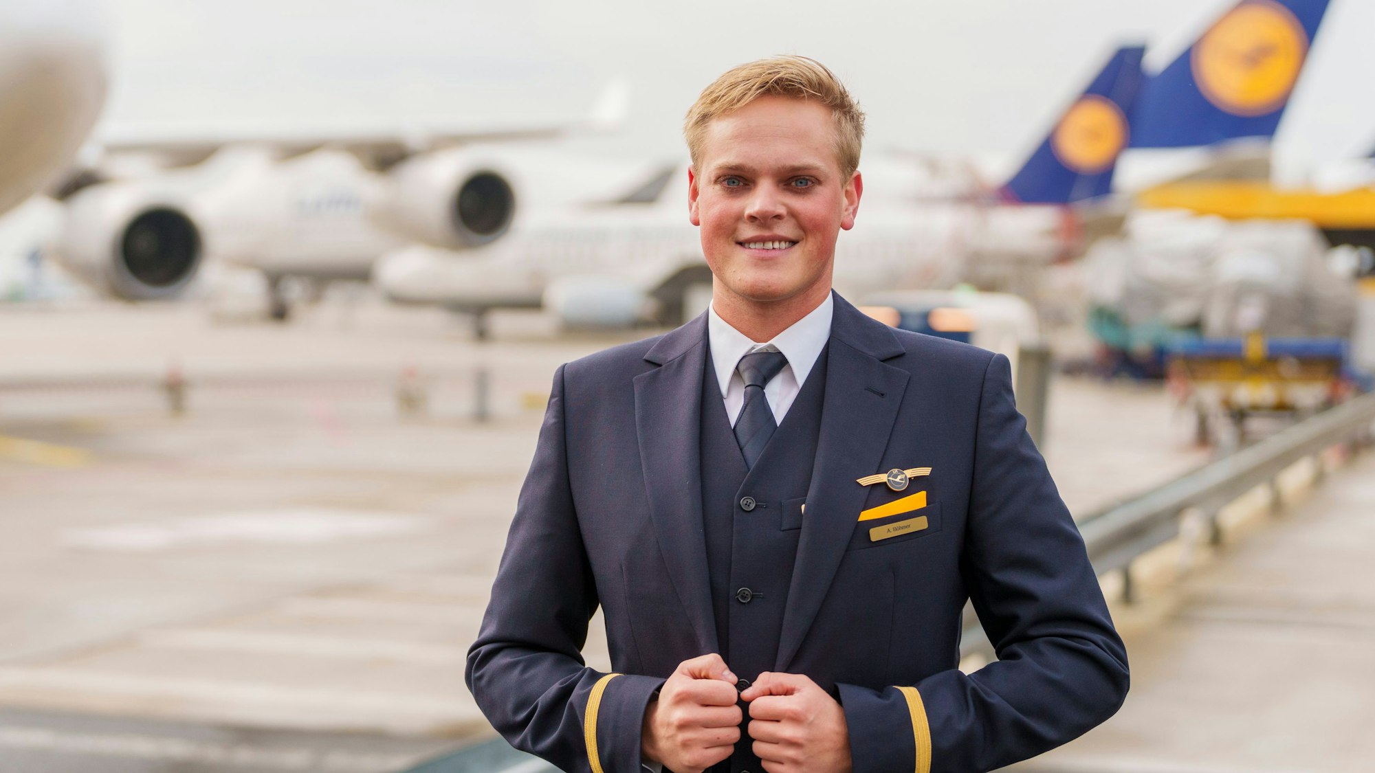 Flugbegleiter Alexander steht in Anzug vor Maschinen der Lufthansa am Flughafen.