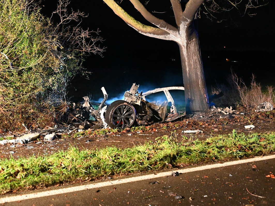 Ein in Teile zerrissenes und ausgebranntes Auto steht an einem Baum.