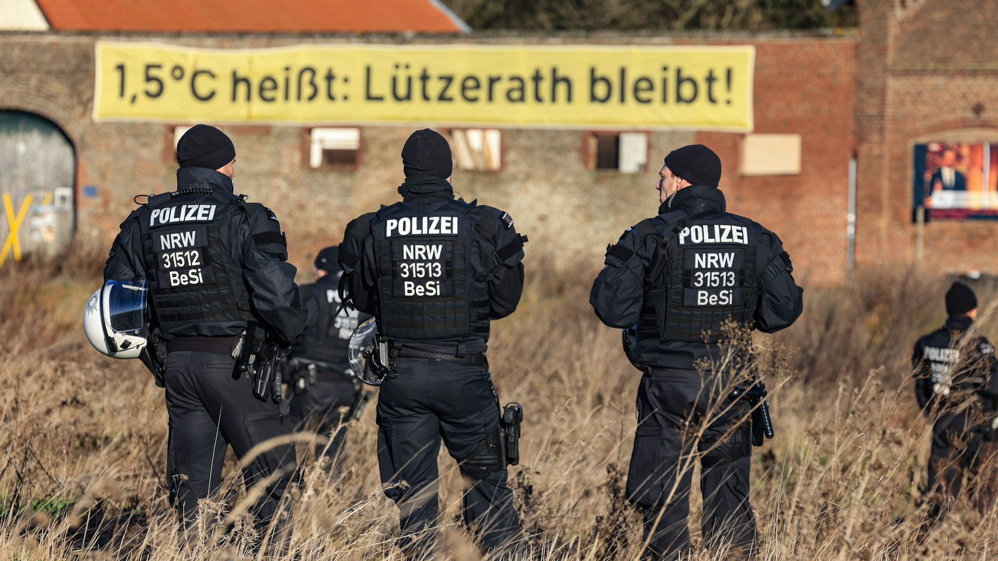 Polizisten stehen in Lützerath am Rande des Braunkohletagebaus, im Hintergrund hängt ein Banner „1,5°C heißt: Lützerath bleibt!“