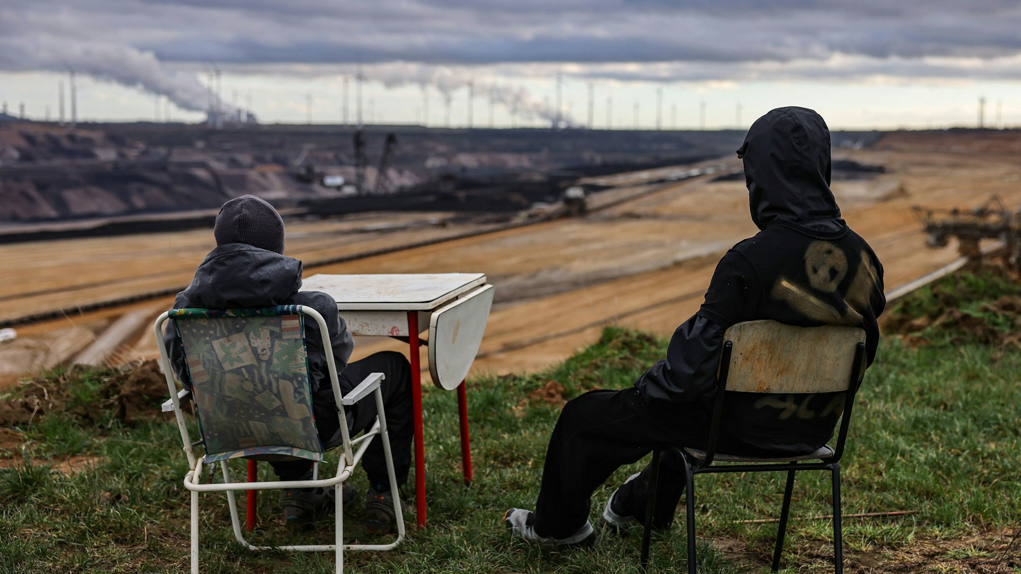 Zwei Personen sitzen am Rande des Braunkohletagebaus auf Stühlen. Sie tragen Kapuze und Mütze. Sie wollen, dass Lützerath bleib.