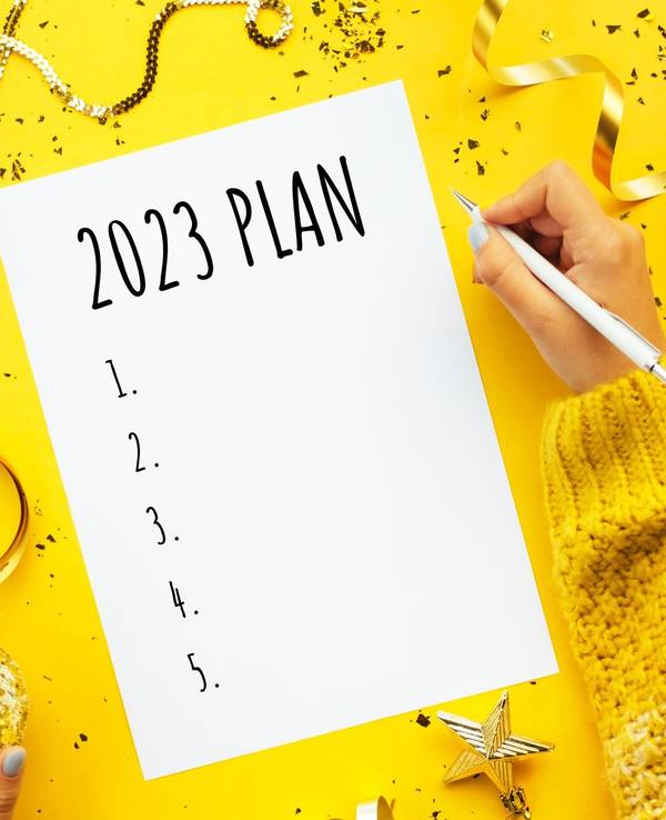 Eine Person schreibt einen Plan für 2023. Weihnachtsdekoration liegt um das Blatt Papier herum auf einem gelben Untergrund. Neben dem Papier ruht die rechte Hand der schreibenden Person mit einem weißen Stift in der Hand.