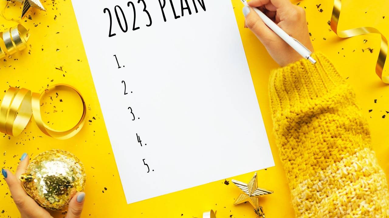 Eine Person schreibt einen Plan für 2023. Weihnachtsdekoration liegt um das Blatt Papier herum auf einem gelben Untergrund. Neben dem Papier ruht die rechte Hand der schreibenden Person mit einem weißen Stift in der Hand.