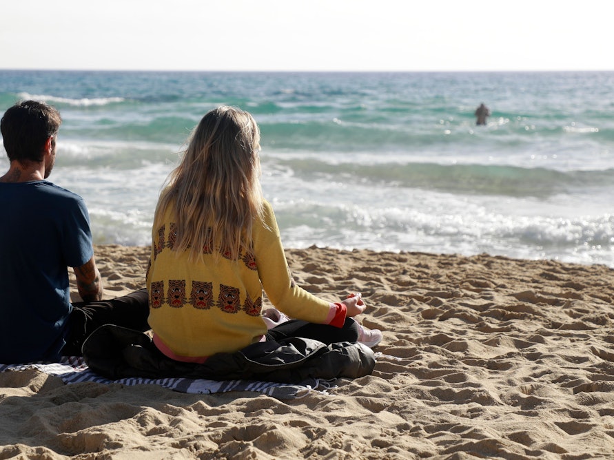 Das Foto zeigt zwei Menschen, die auf einer Decke am Strand sitzen und auf das Meer schauen.