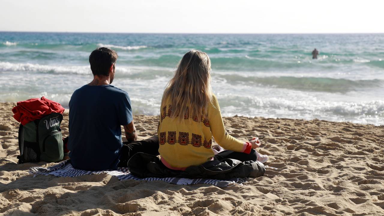 Das Foto zeigt zwei Menschen, die auf einer Decke am Strand sitzen und auf das Meer schauen.
