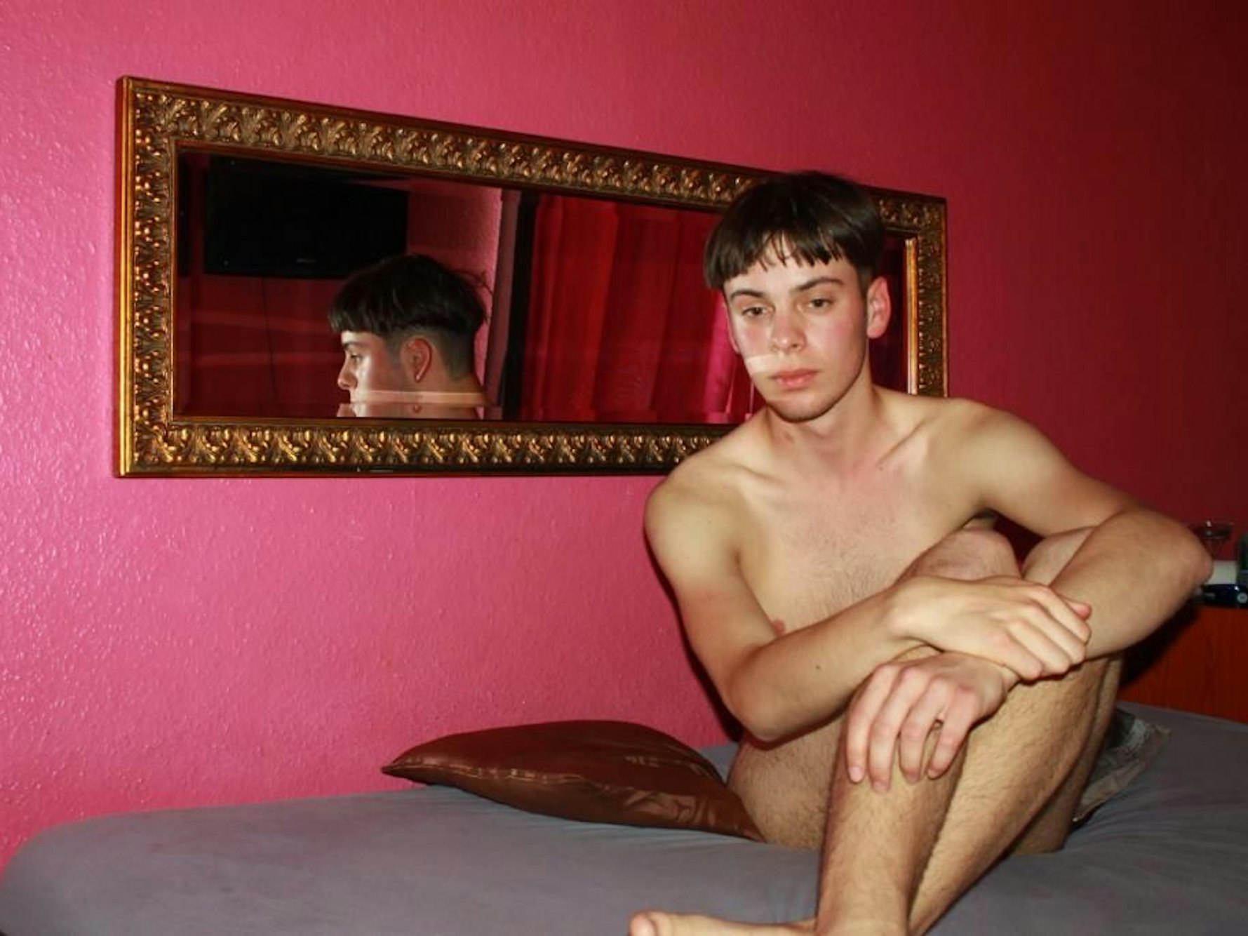 Alex sitzt nackt auf einem Bett.