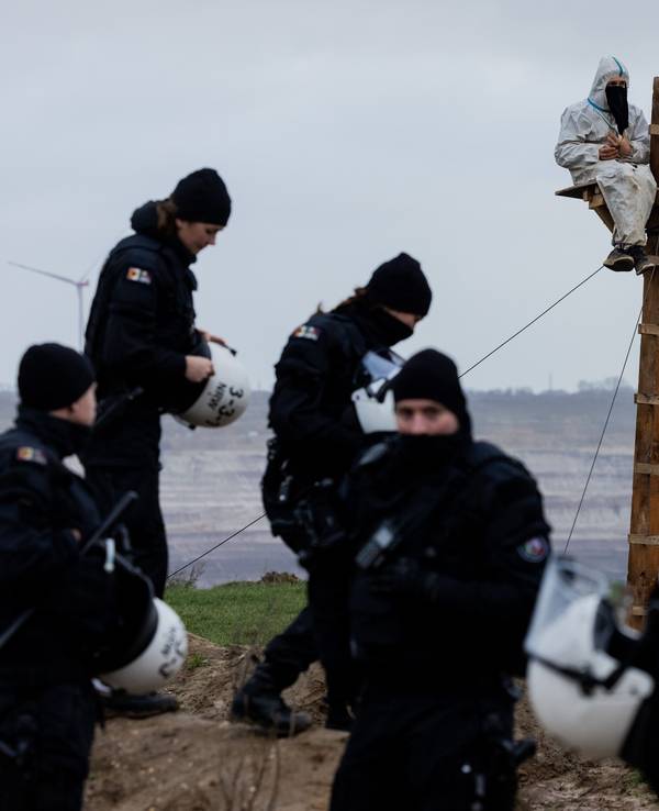 Ein Umweltaktivist sitzt auf einem selbstgebauten Hochsitz unmittelbar an der Abrisskante des Braunkohletagebaus Garzweiler II. Polizeikräfte stehen drumherum.