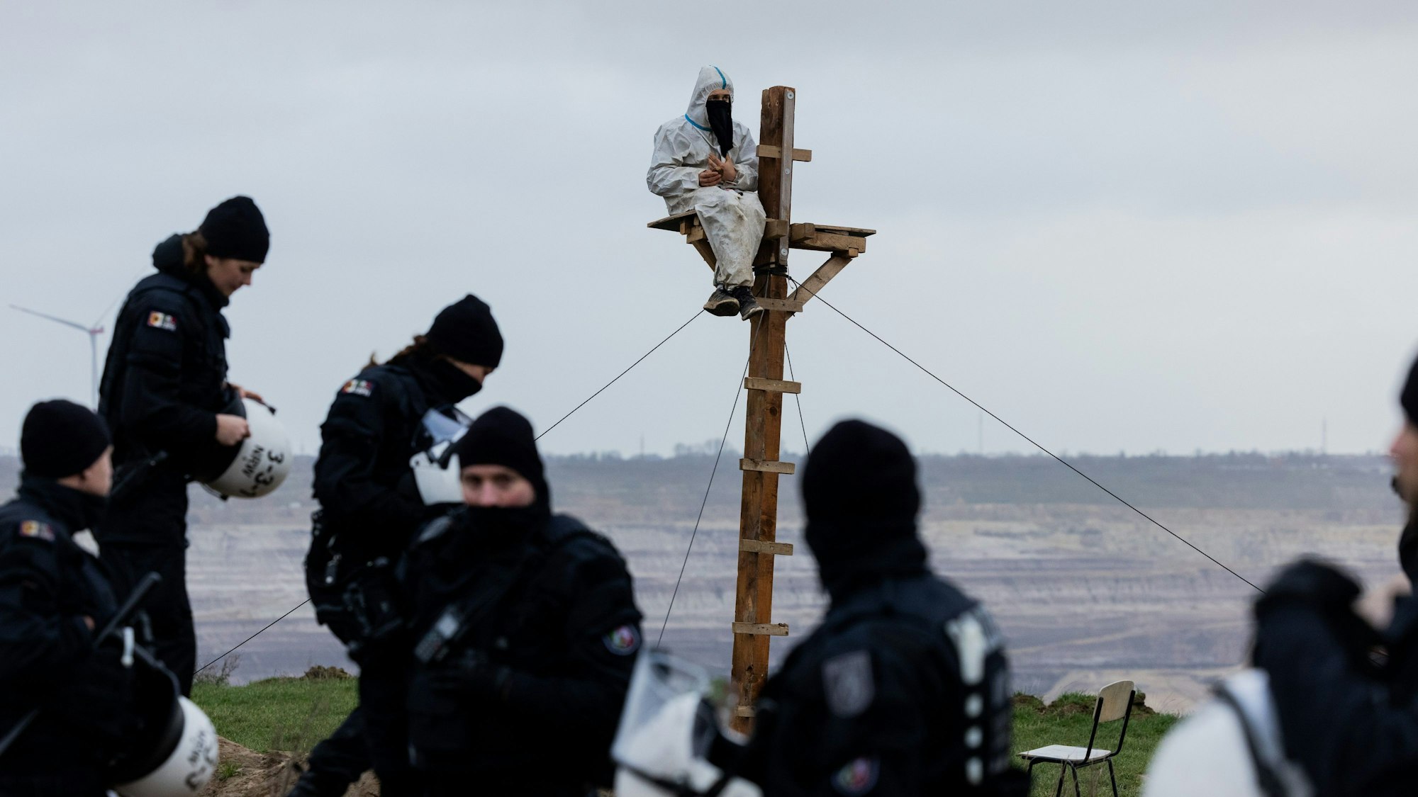 Ein Umweltaktivist sitzt auf einem selbstgebauten Hochsitz unmittelbar an der Abrisskante des Braunkohletagebaus Garzweiler II. Polizeikräfte stehen drumherum.