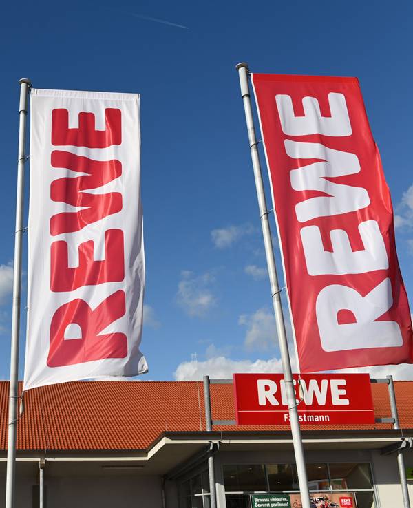 Fahnen mit dem Logo des Lebensmitteleinzelhändlers Rewe wehen vor einer Filiale.