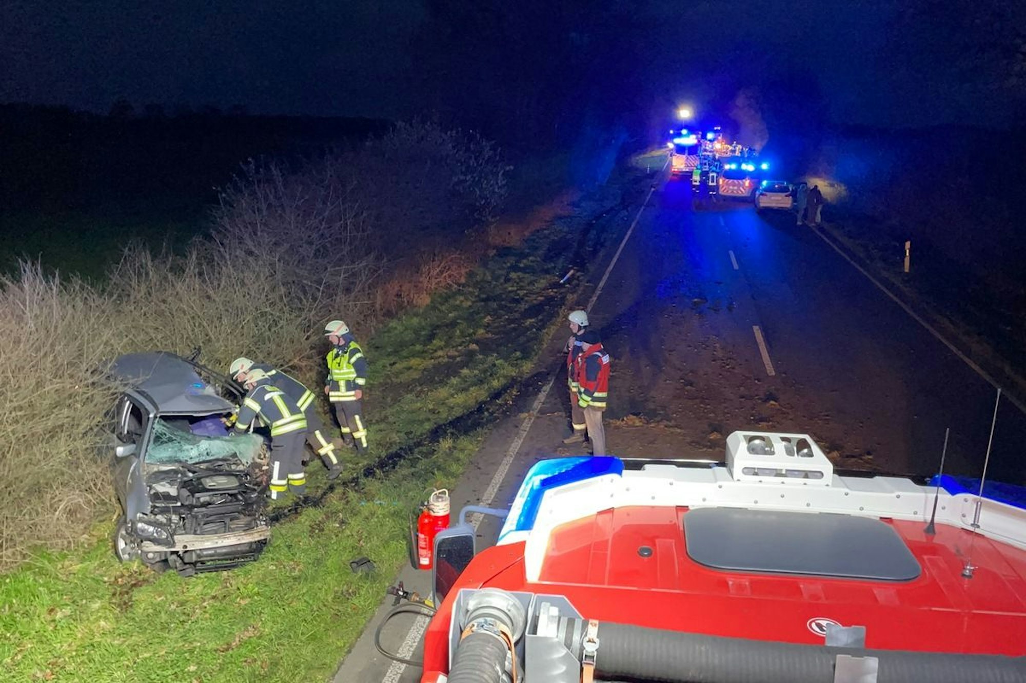 Ein Auto steht schwer beschädigt neben der Straße, mehrere Einsatzkräfte stehen davor. Auf der Straße befinden sich mehrere Einsatzfahrzeuge.