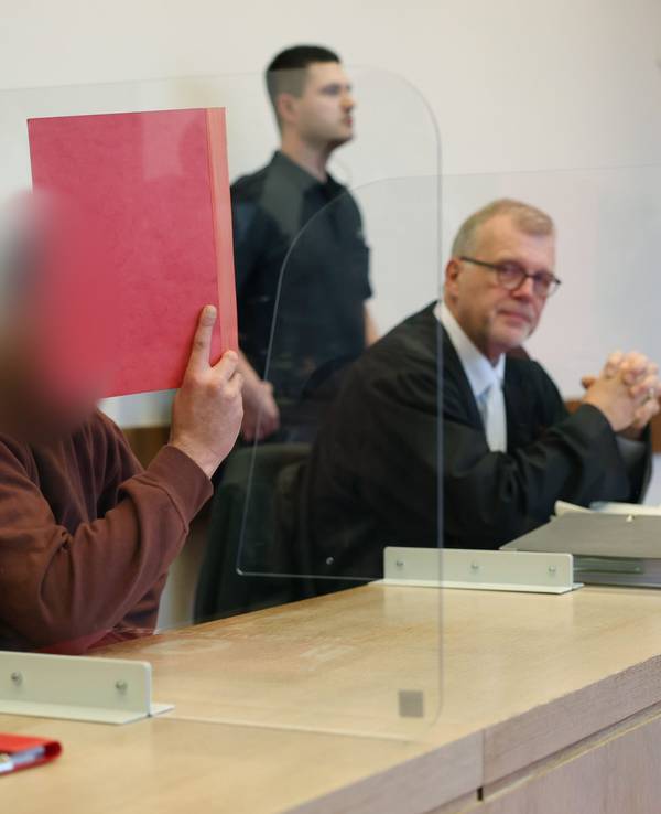 Der mutmaßliche Axtmörder sitzt neben seinem Verteidiger auf der Anklagebank und hält sich eine Mappe vor das Gesicht.