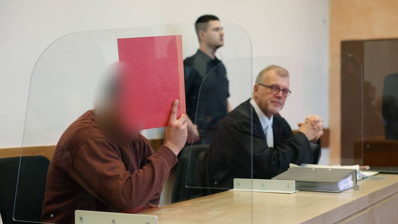 Der mutmaßliche Axtmörder sitzt neben seinem Verteidiger auf der Anklagebank und hält sich eine Mappe vor das Gesicht.