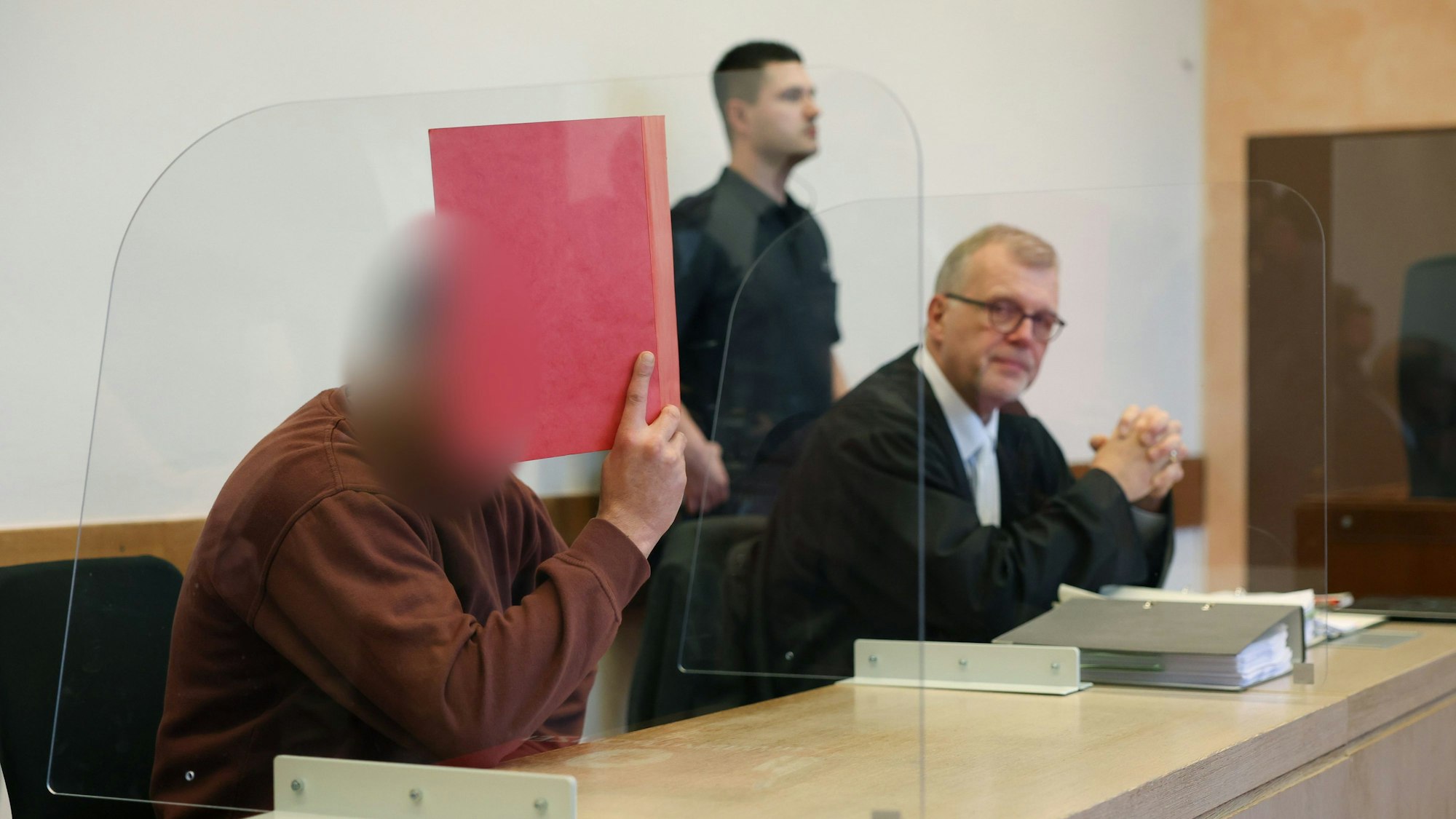 Der mutmaßliche Axtmörder sitzt neben seinem Verteidiger auf der Anklagebank und hält sich eine Mappe vor das Gesicht.