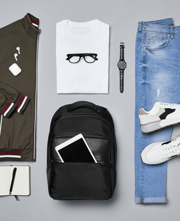 Verschiedene Kleidungsstücke und Accessoires wie eine Jacke, ein T-Shirt, eine Armbanduhr, ein Paar Schuhe, eine Jeans, ein Rucksack und ein iPad, ein Notizblock und ein Kugelschreiber liegen auf einem grauen Untergrund.