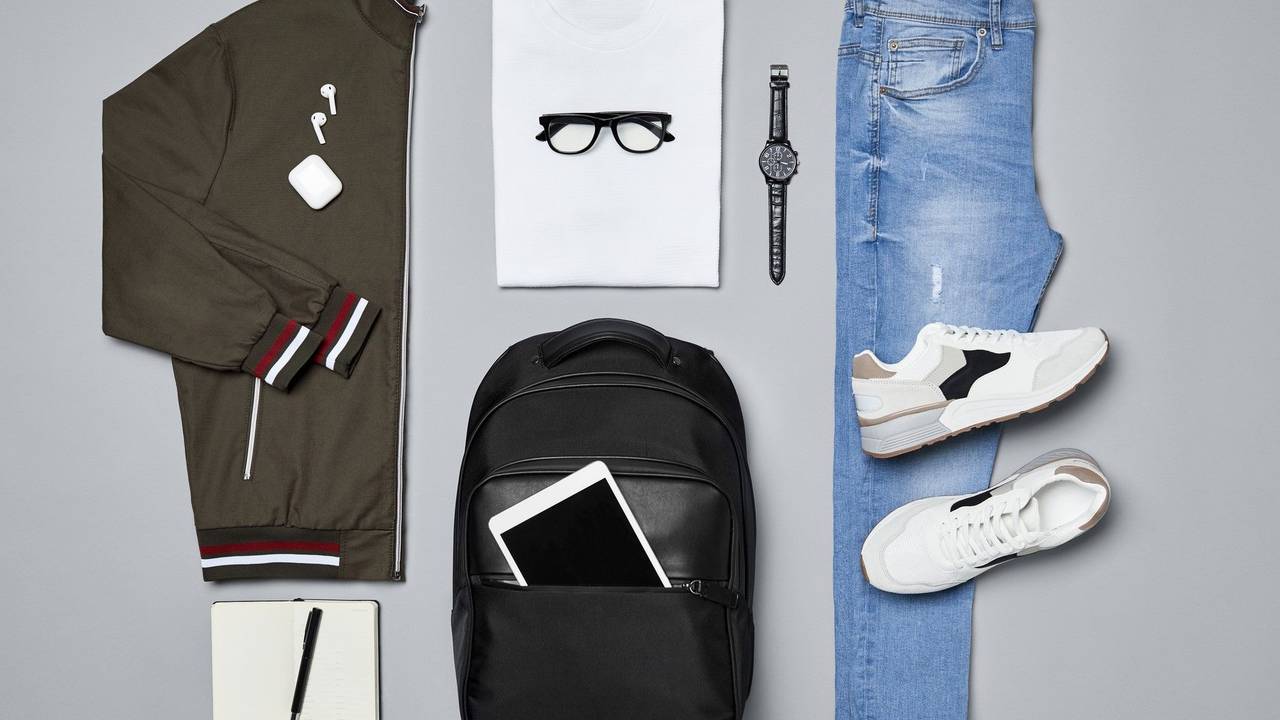 Verschiedene Kleidungsstücke und Accessoires wie eine Jacke, ein T-Shirt, eine Armbanduhr, ein Paar Schuhe, eine Jeans, ein Rucksack und ein iPad, ein Notizblock und ein Kugelschreiber liegen auf einem grauen Untergrund.