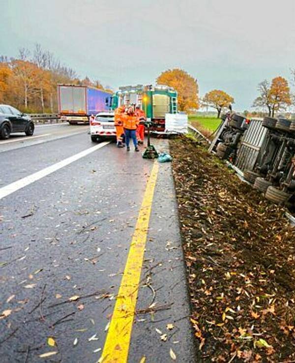 Ein Lkw liegt umgekippt neben der Fahrbahn der Autobahn. Einsatzkräfte stehen daneben.