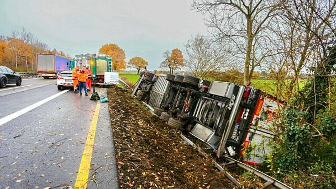 Ein Lkw liegt umgekippt neben der Fahrbahn der Autobahn. Einsatzkräfte stehen daneben.