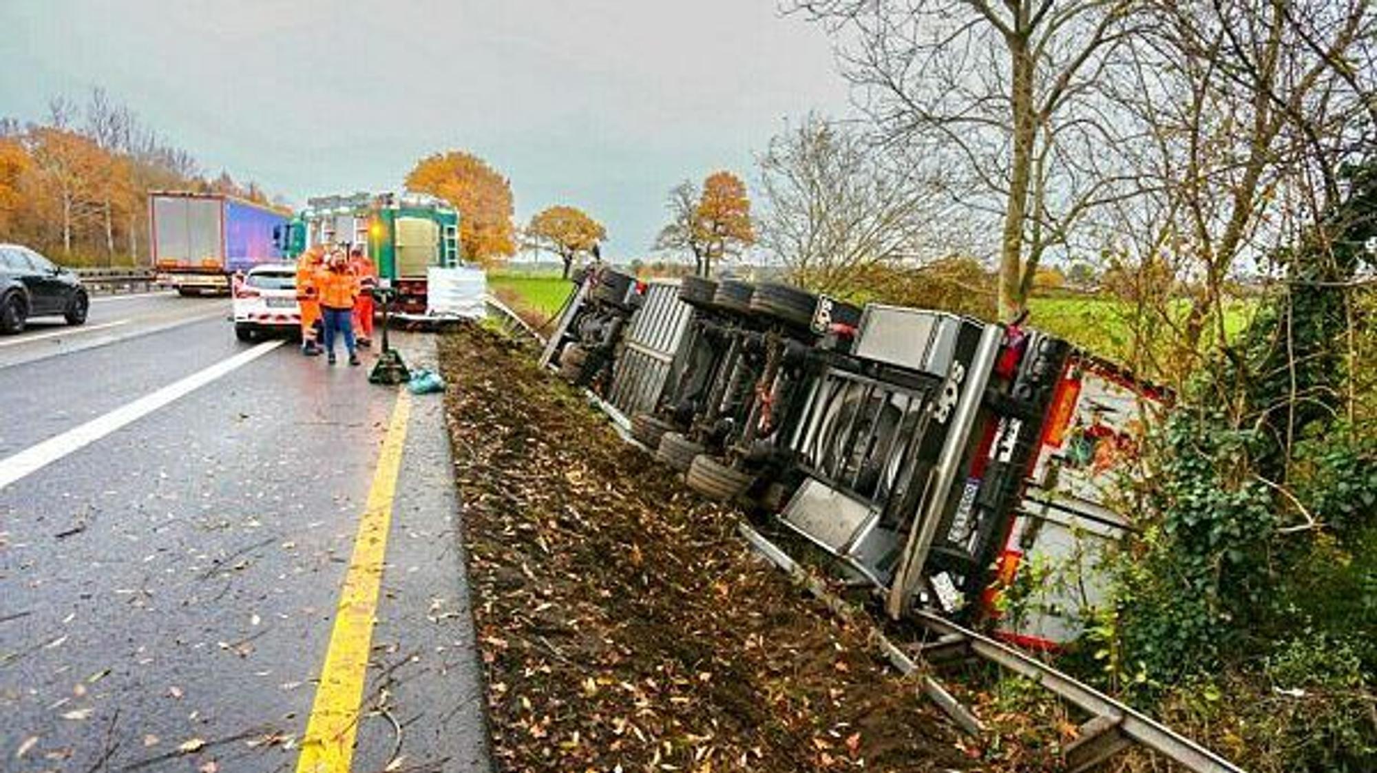 Ein Lkw liegt umgekippt neben der Fahrbahn der Autobahn. Einsatzkräfte stehen daneben.