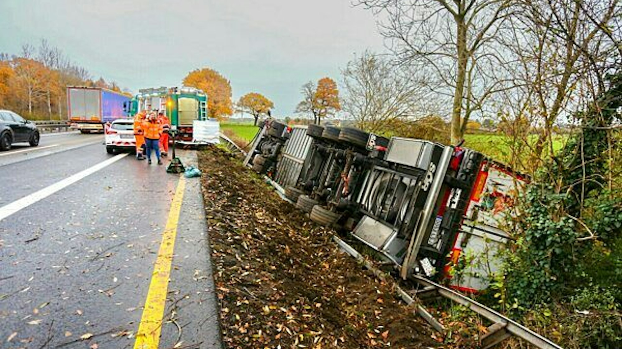 Ein Lkw liegt umgekippt neben der Fahrbahn der Autobahn. Einsatzkräfte stehen daneben.