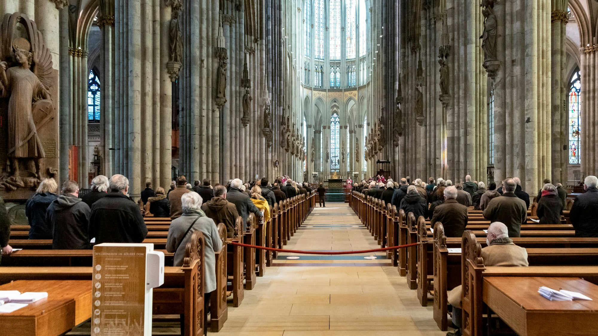 Menschen sitzen in den Bänken des Kölner Doms. Der Blick vom Foto aus geht genau mittig durch die Reihen auf den Altar.