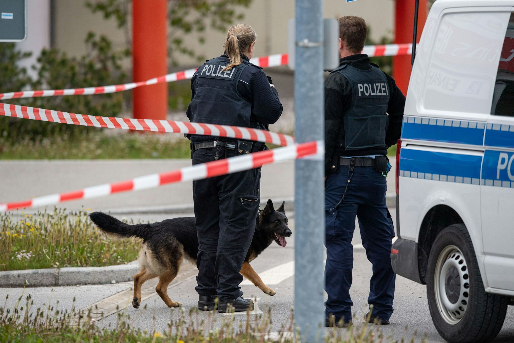 Zwei Polizisten und ein Spürhund stehen vor einem abgesperrten Bereich.