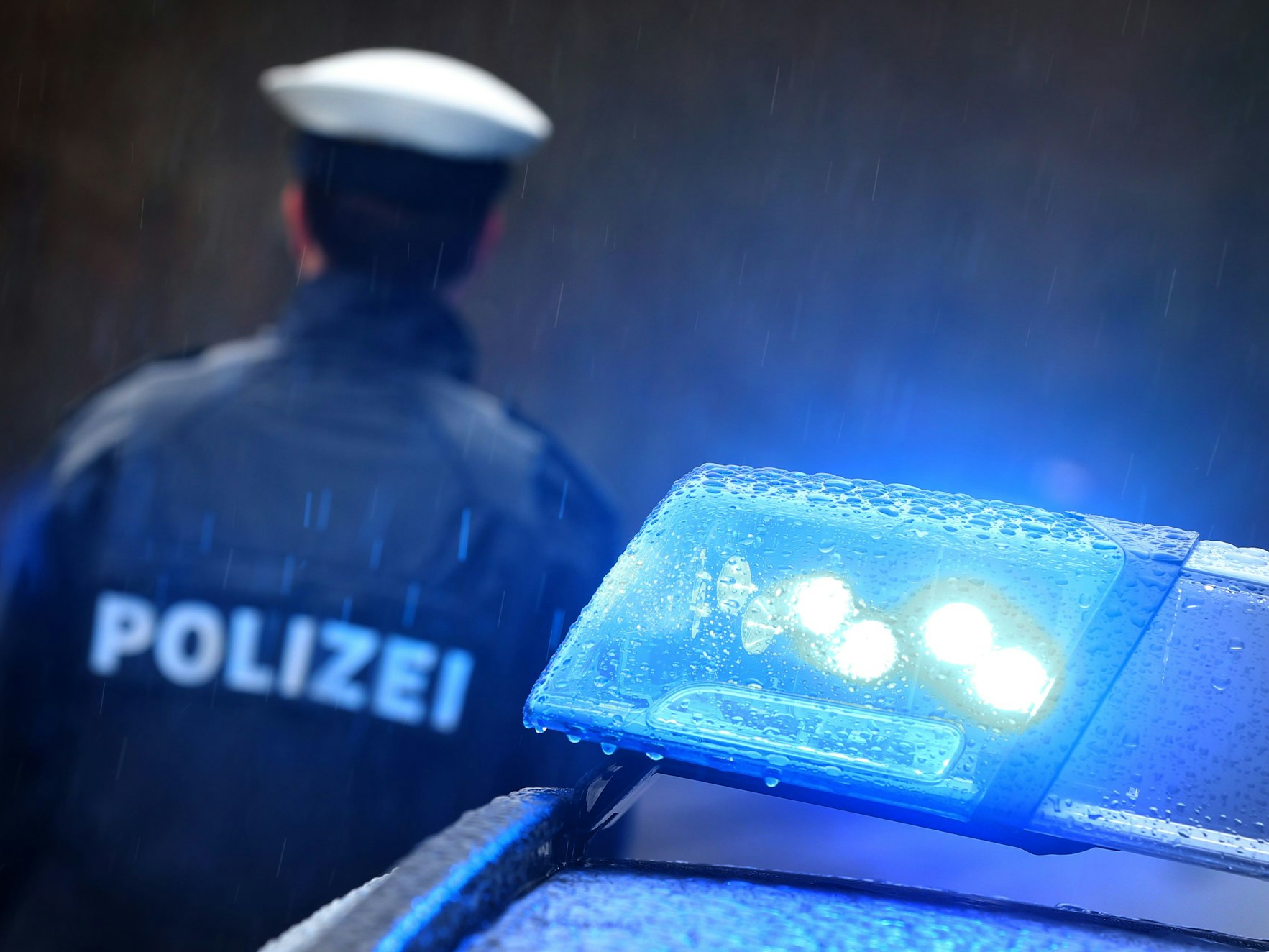 Ein Polizist steht im Regen vor einem Streifenwagen dessen Blaulicht aktiviert ist.