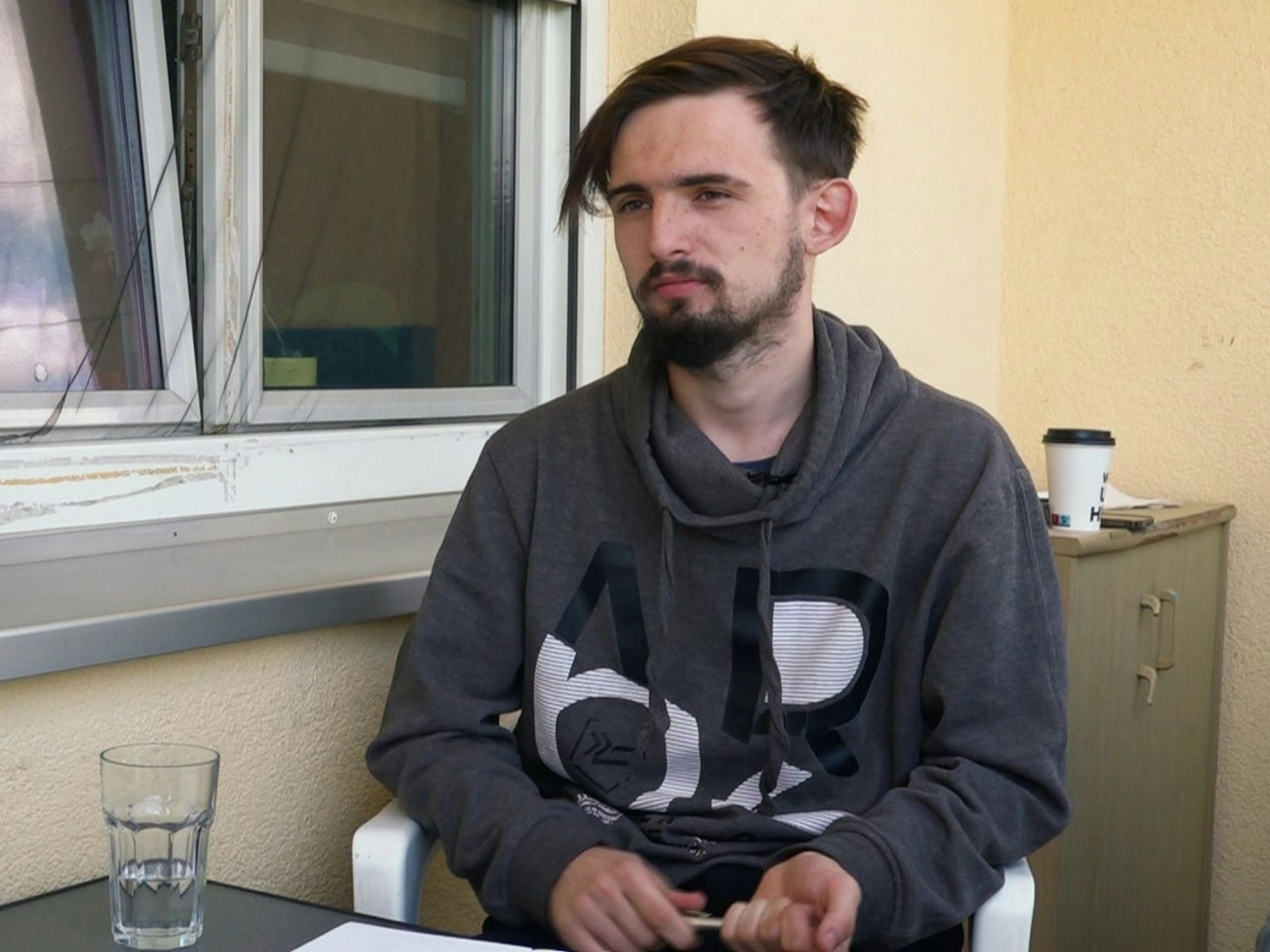 Jerrick sitzt auf einem Balkon. Er hält einen Stift in der Hand, vor ihm liegt ein Blatt Papier.