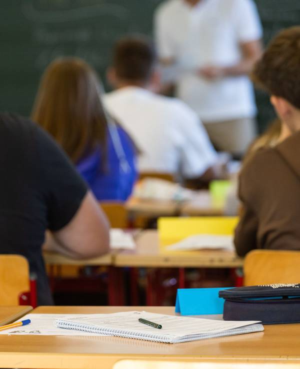 Arbeitsmaterialien liegen auf einem Tisch in einem Klassenzimmer während dahinter Schüler, Schülerinnen und ihr Lehrer zu sehen sind.