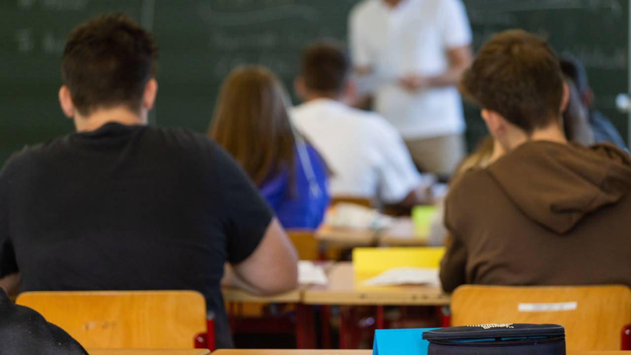 Arbeitsmaterialien liegen auf einem Tisch in einem Klassenzimmer während dahinter Schüler, Schülerinnen und ihr Lehrer zu sehen sind.