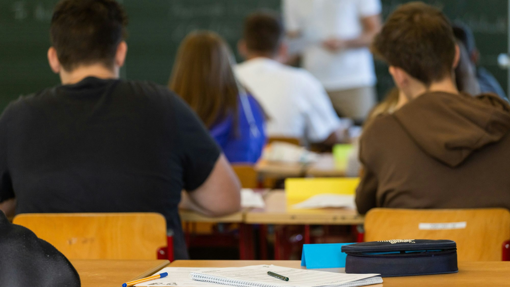Arbeitsmaterialien liegen auf einem Tisch in einem Klassenzimmer während dahinter Schüler, Schülerinnen und ihr Lehrer zu sehen sind.