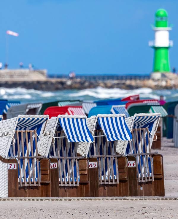 Das Symbolfoto zeigt leere Strandkörbe vor blauem Meer, blauem Himmel und einem weiß-grünen Leuchtturm.
