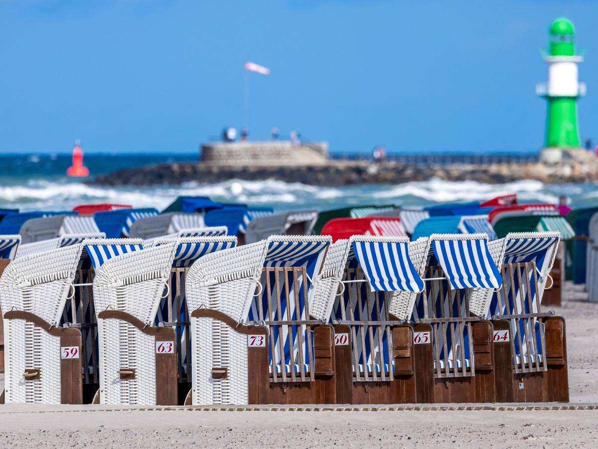 Das Symbolfoto zeigt leere Strandkörbe vor blauem Meer, blauem Himmel und einem weiß-grünen Leuchtturm.