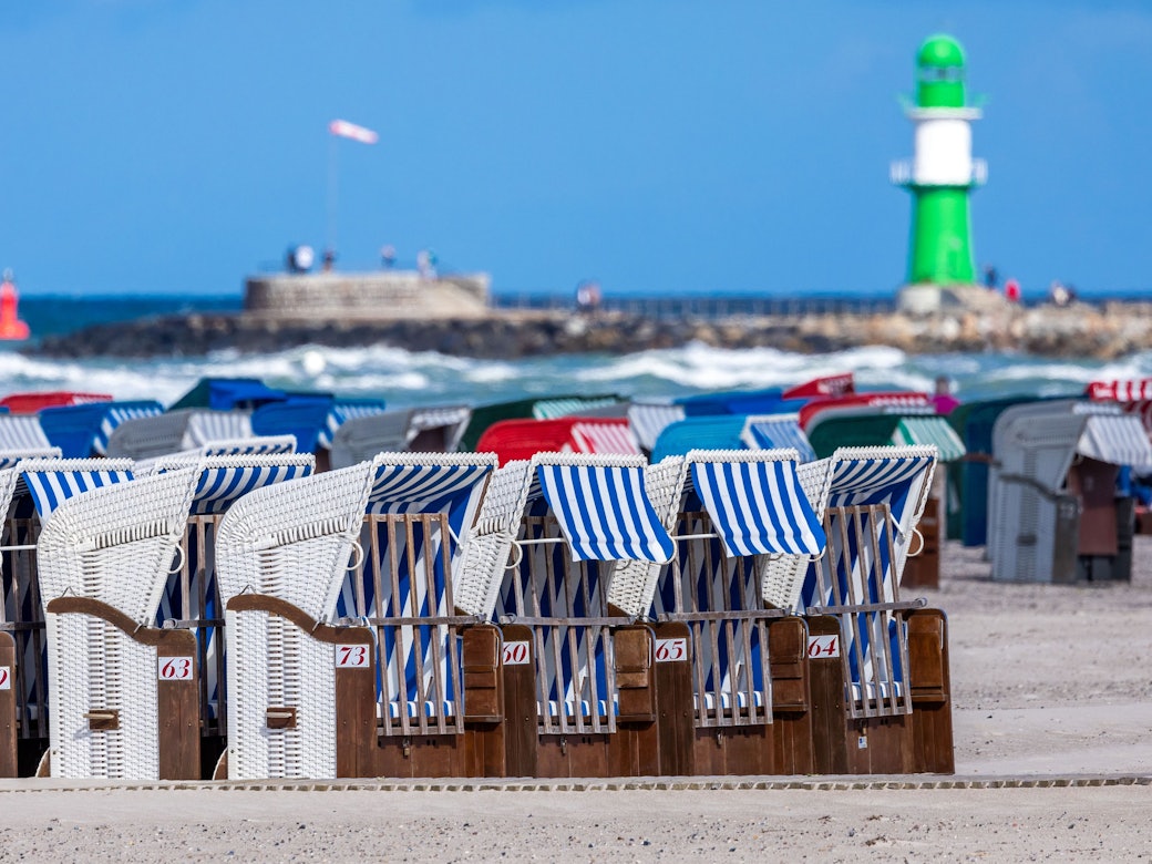 Das Symbolfoto zeigt leere Strandkörbe vor blauem Meer, blauem Himmel und einem weiß-grünen Leuchtturm.