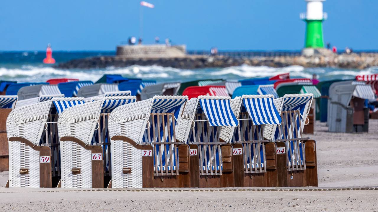 Das Symbolfoto zeigt leere Strandkörbe vor blauem Meer, blauem Himmel und einem weiß-grünen Leuchtturm.
