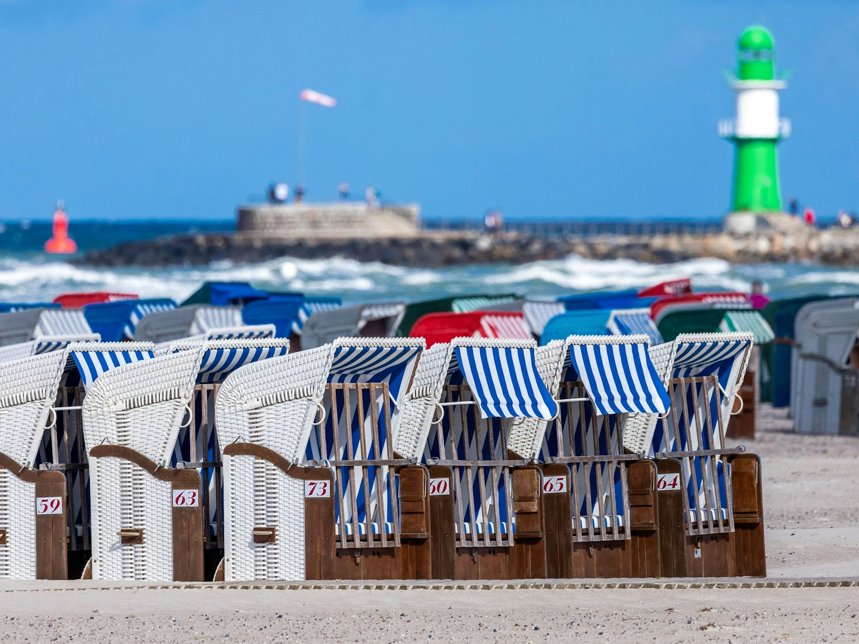 Das Symbolfoto zeigt leere Strandkörbe vor blauem Meer, blauem Himmel und einem weiß-grünen Leuchtturm.