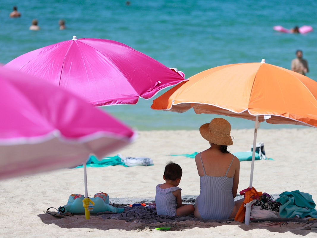 Eine Frau sitzt neben ihrem Kind unter einem Sonnenschirm an einem warmen Sommertag am Strand von Mallorca.