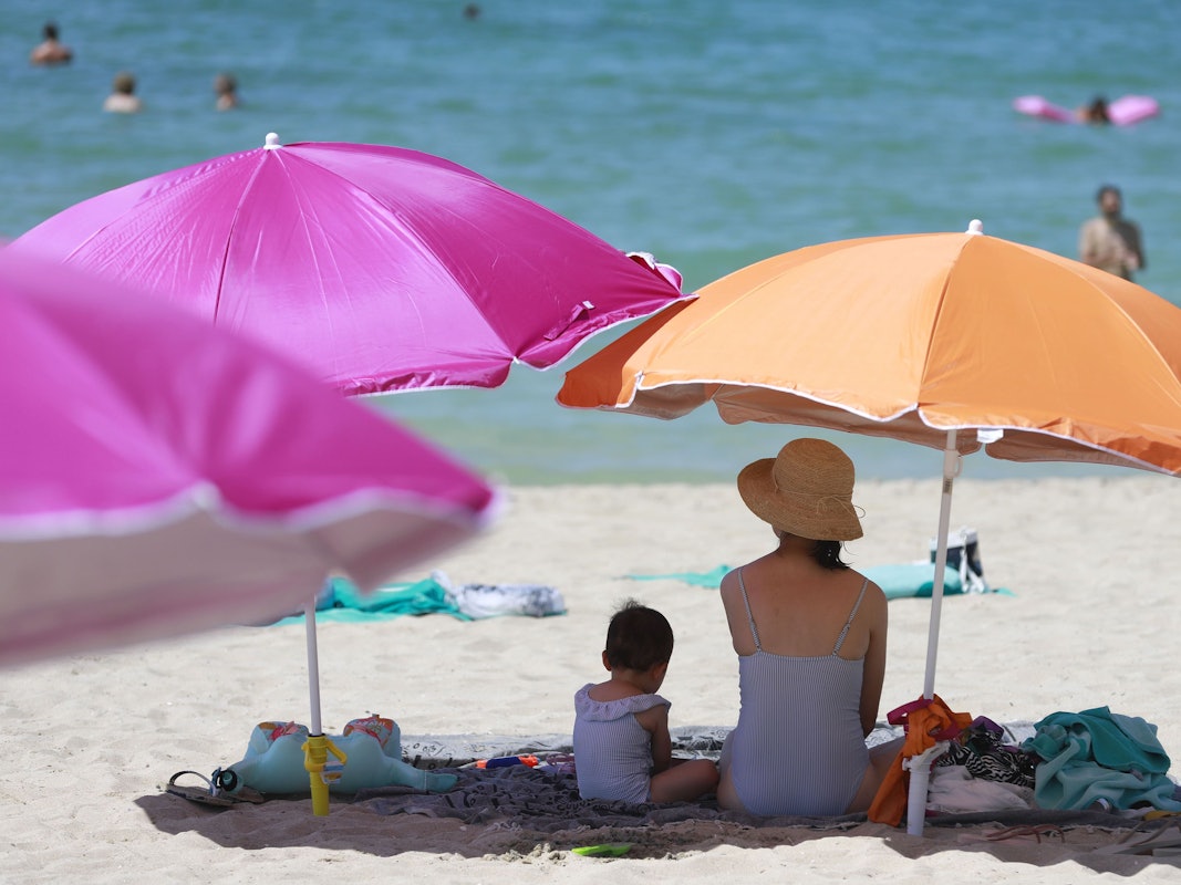 Eine Frau sitzt neben ihrem Kind unter einem Sonnenschirm an einem warmen Sommertag am Strand von Palma de Mallorca im August 2022. Mallorca bleibt das günstigste Reiseziel für Familien im Jahr 2023. Doch einen Haken hat die ganz Sache trotzdem.